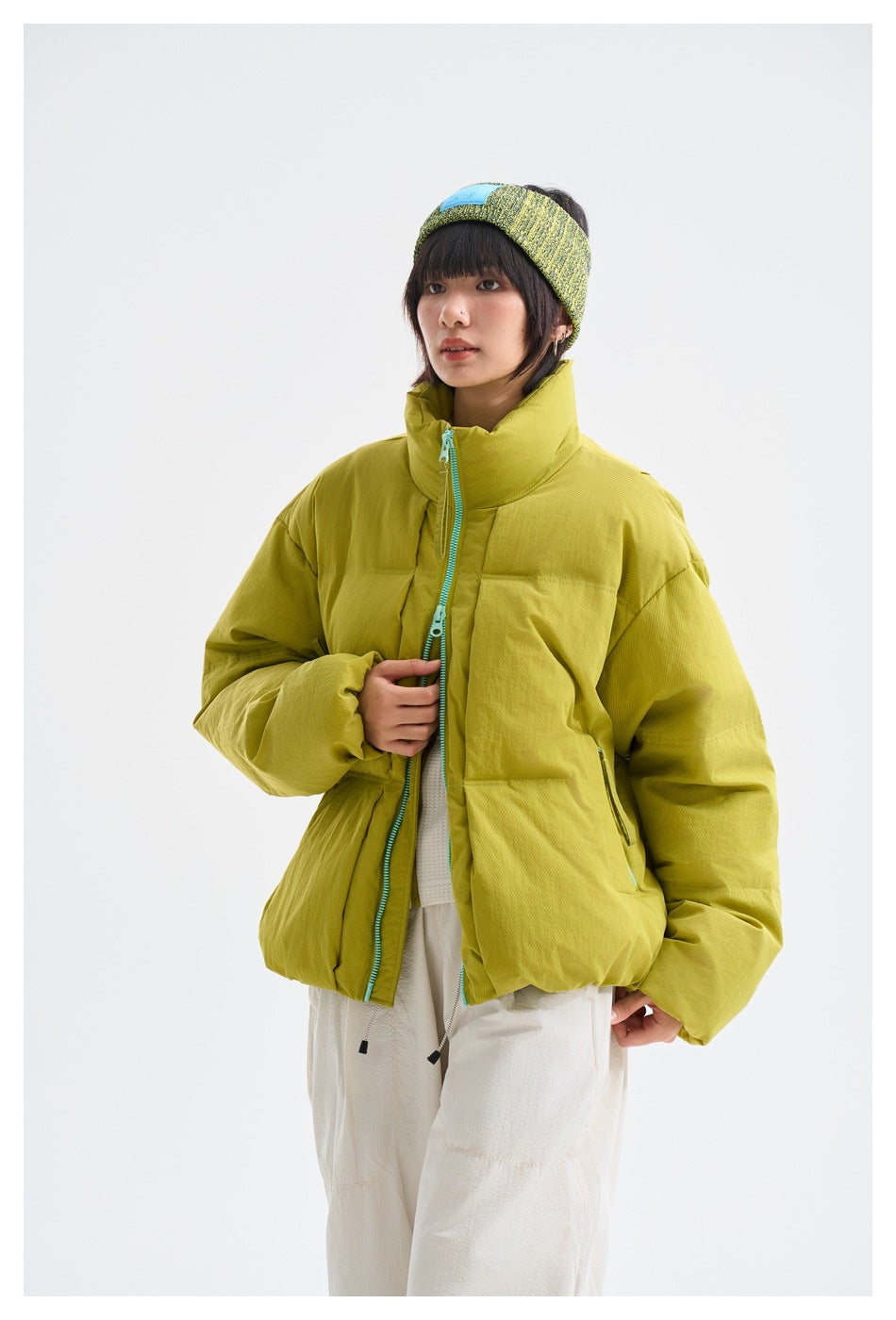 Model wearing a chartreuse oversized puffer jacket with turquoise zipper, white trousers, and a green-yellow knit headband.