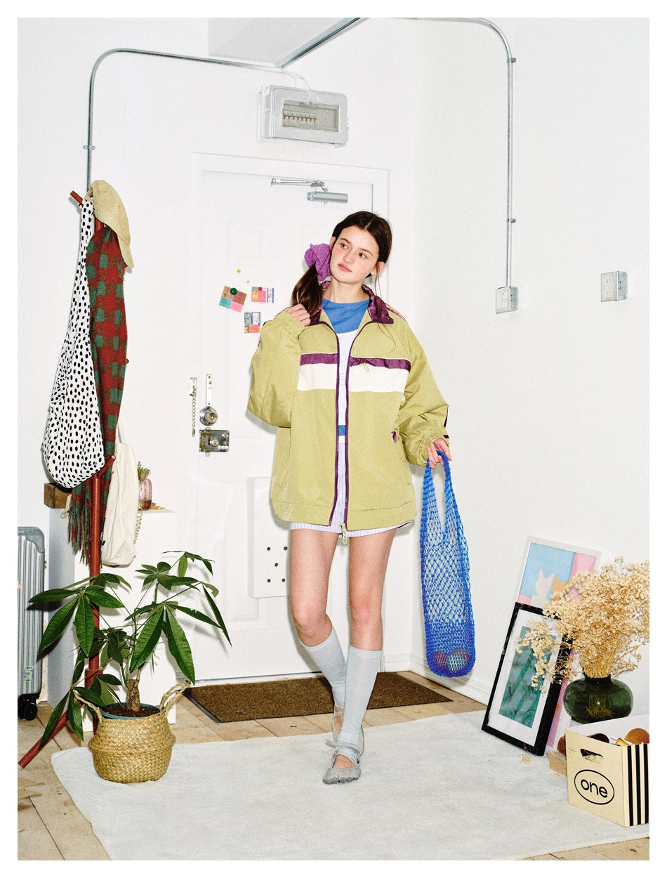 Model poses in a khaki track jacket with purple-and-white stripes, holding a blue mesh bag.