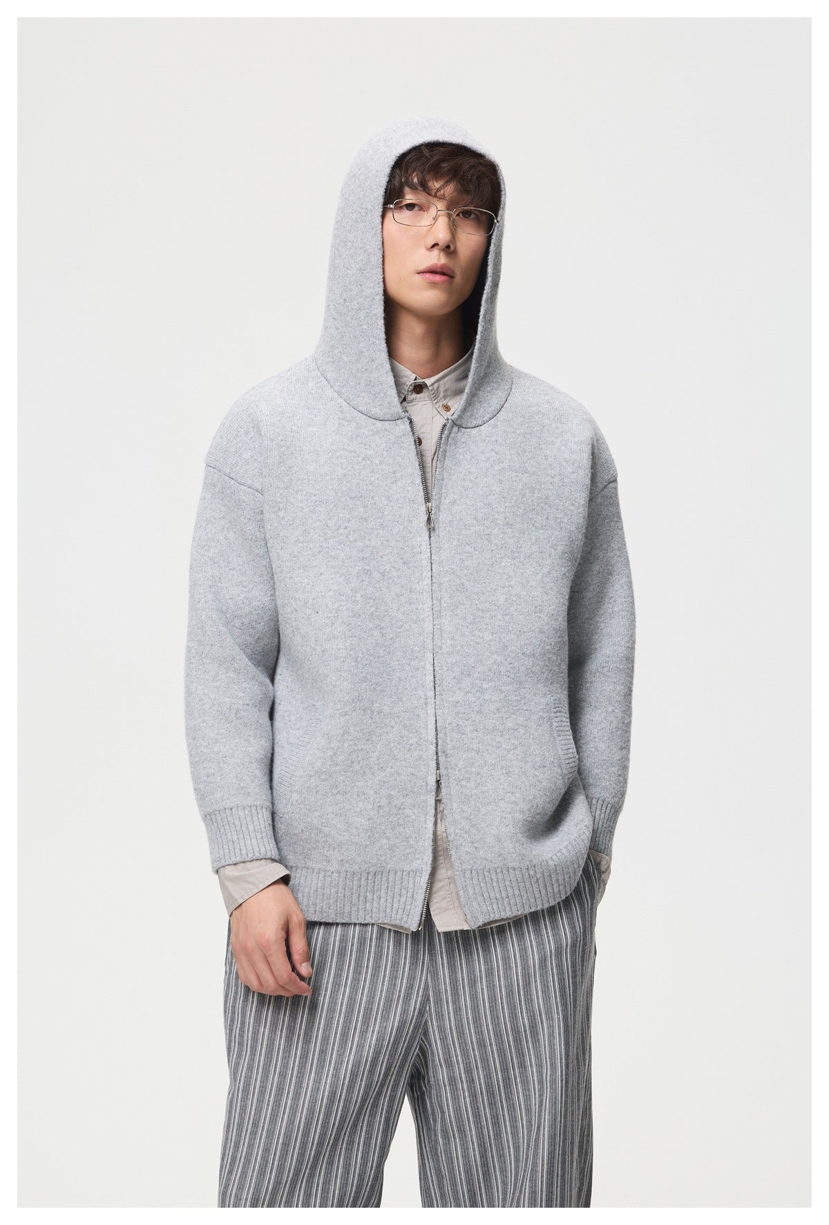 Model wearing the light gray knit zip hooded jacket with the hood up over a beige shirt and striped trousers