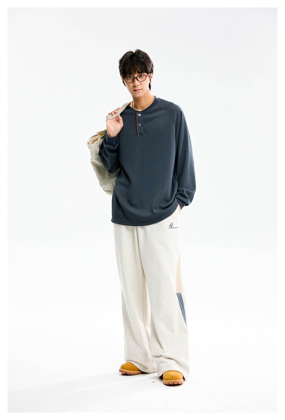 Model wearing a slate-blue long-sleeve Henley shirt with cream pants, carrying a light-colored shoulder bag.