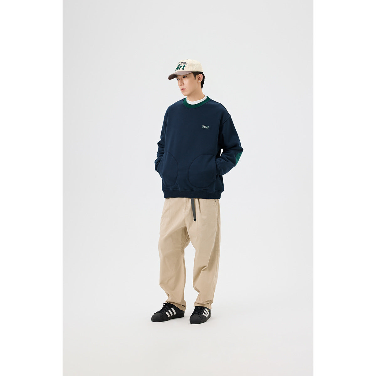 Model in a navy sweatshirt with green collar trim and rounded front pockets, beige trousers, and a cream cap.