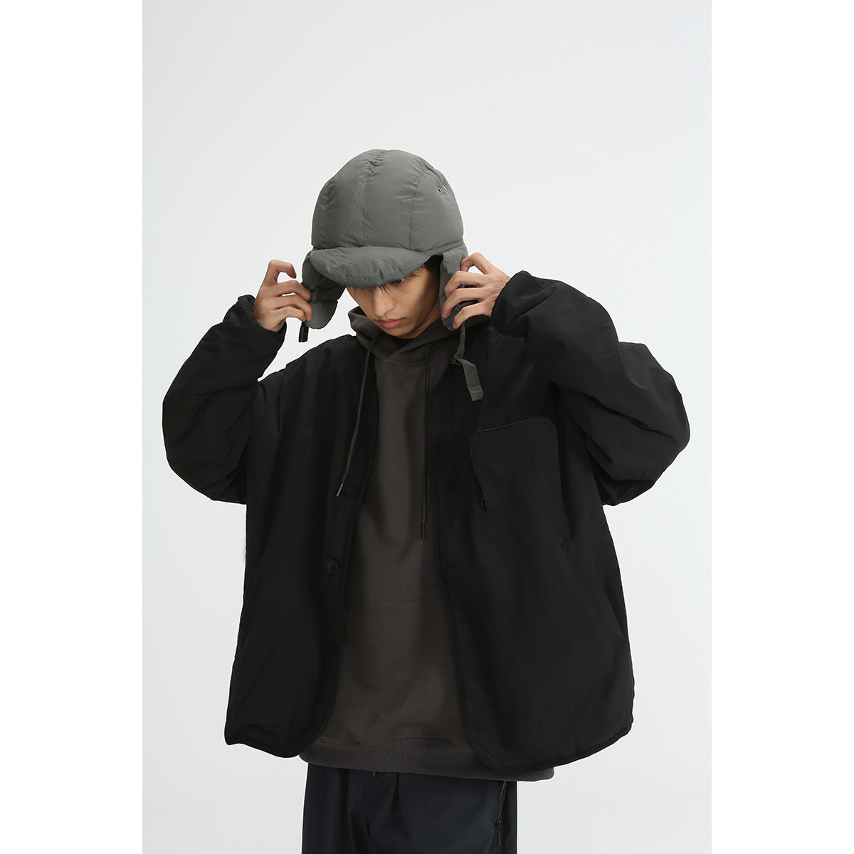 Model wearing a roomy black button-front jacket with a chest pocket, layered over a dark hoodie, and a gray earflap cap.