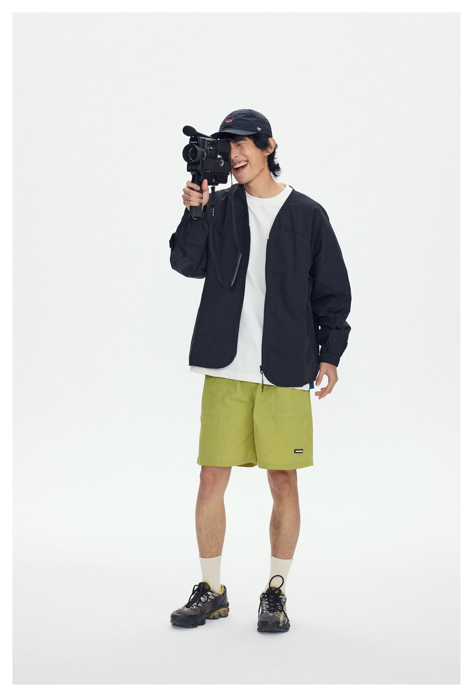 Model wearing a black open jacket, white shirt and lime green shorts while holding a video camera.