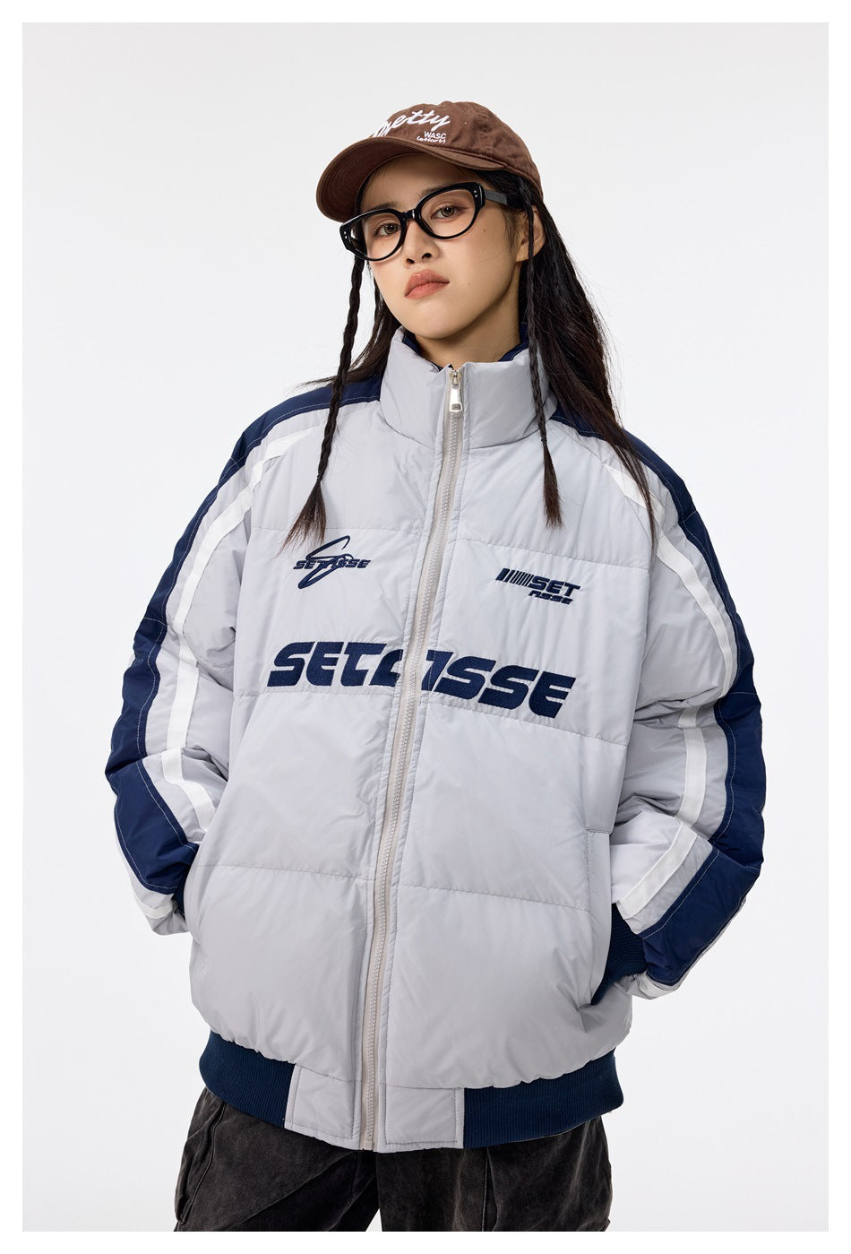 Model wearing a light gray padded zip jacket with navy and white sleeve stripes, dark ribbed trim, and navy chest graphics.