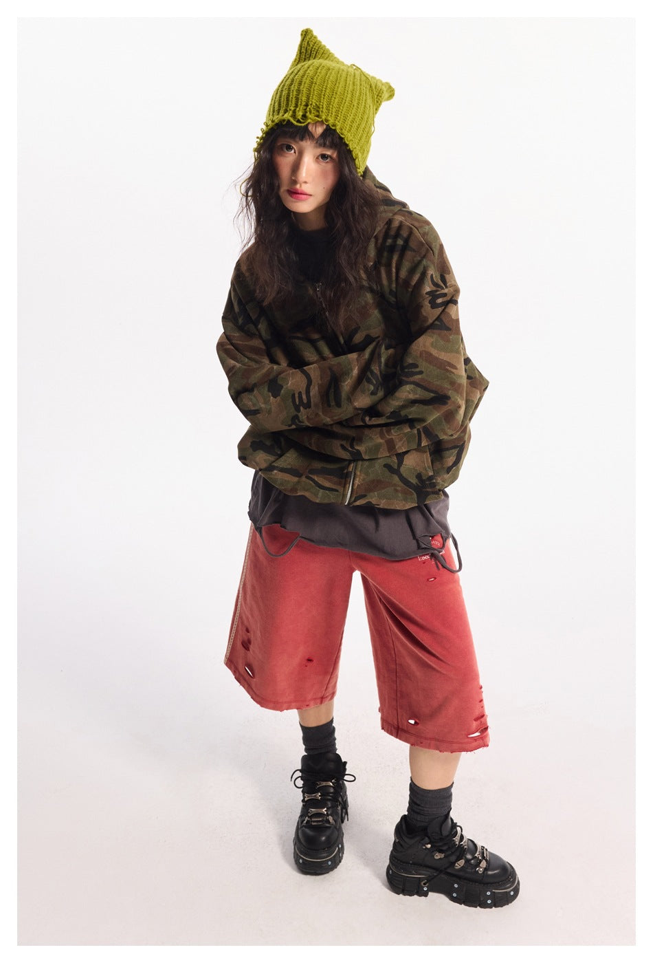 Model in a camouflage zip jacket and distressed red wide-leg shorts, wearing a lime knit beanie and black platform shoes.