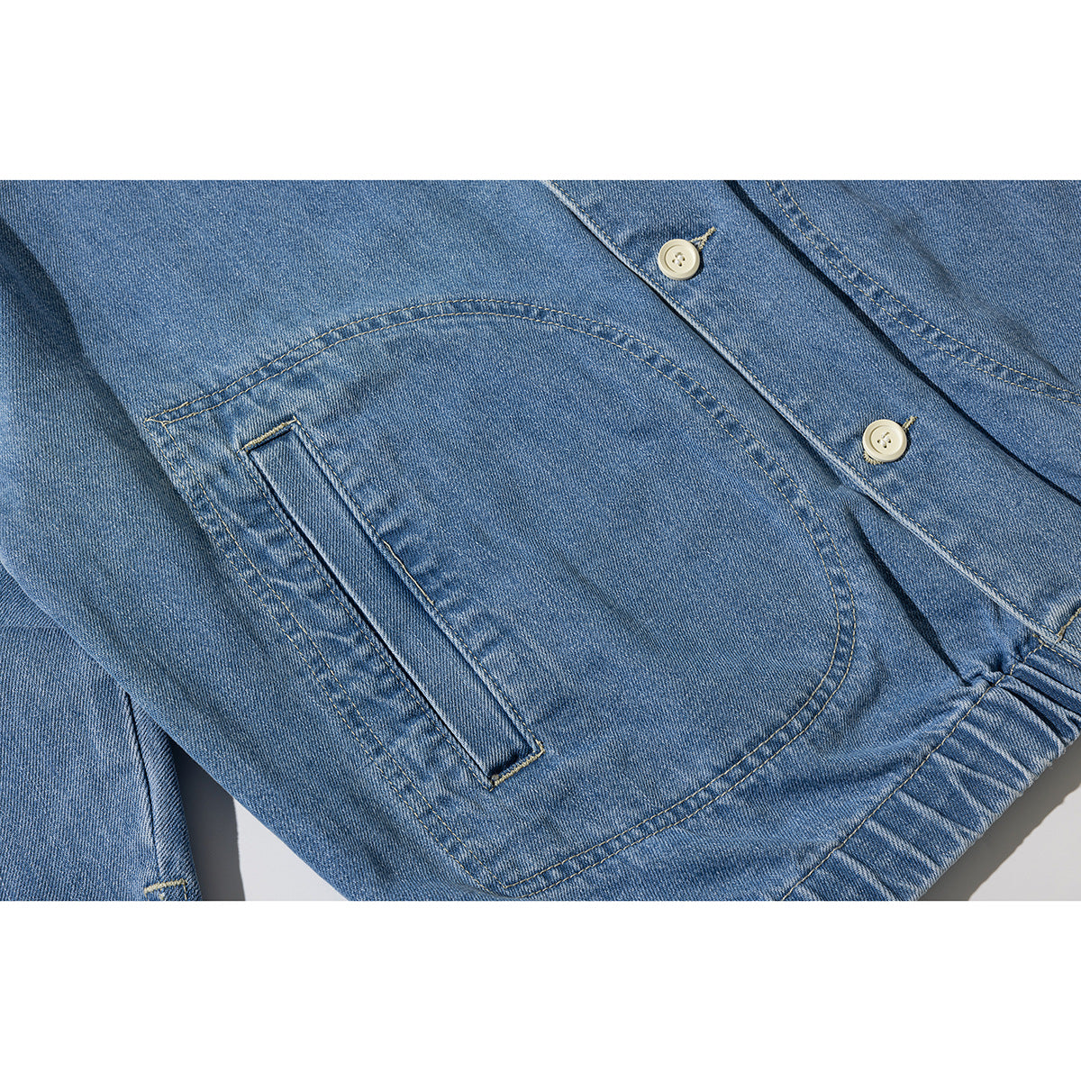 Close-up of a blue denim jacket's curved front patch pocket, cream buttons, and contrast stitching.