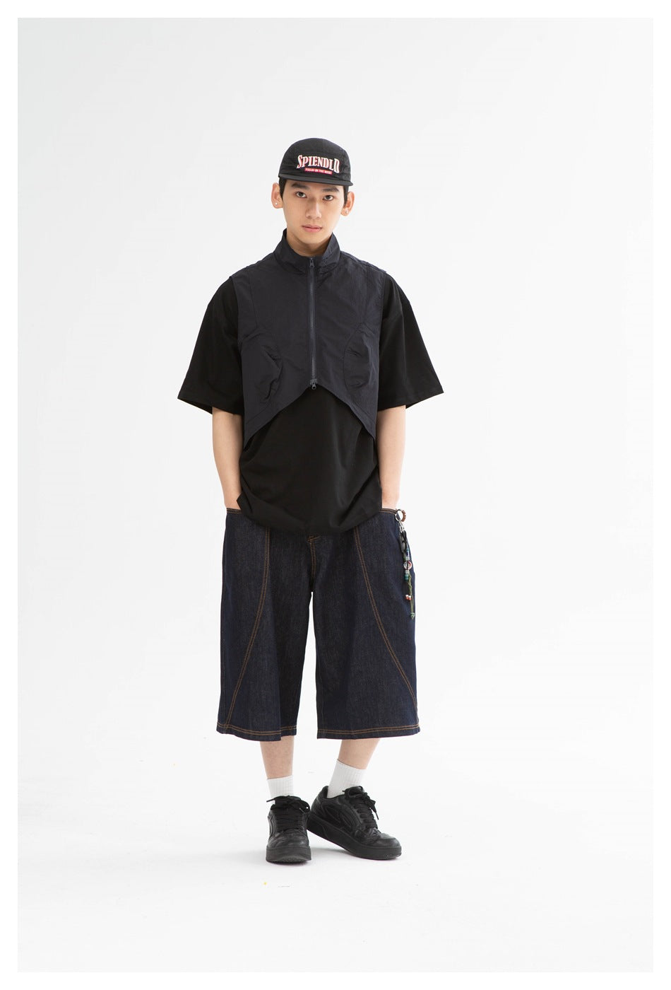 Model wearing dark indigo wide-leg denim shorts with contrast stitching, a black shirt, cropped vest, cap and sneakers.