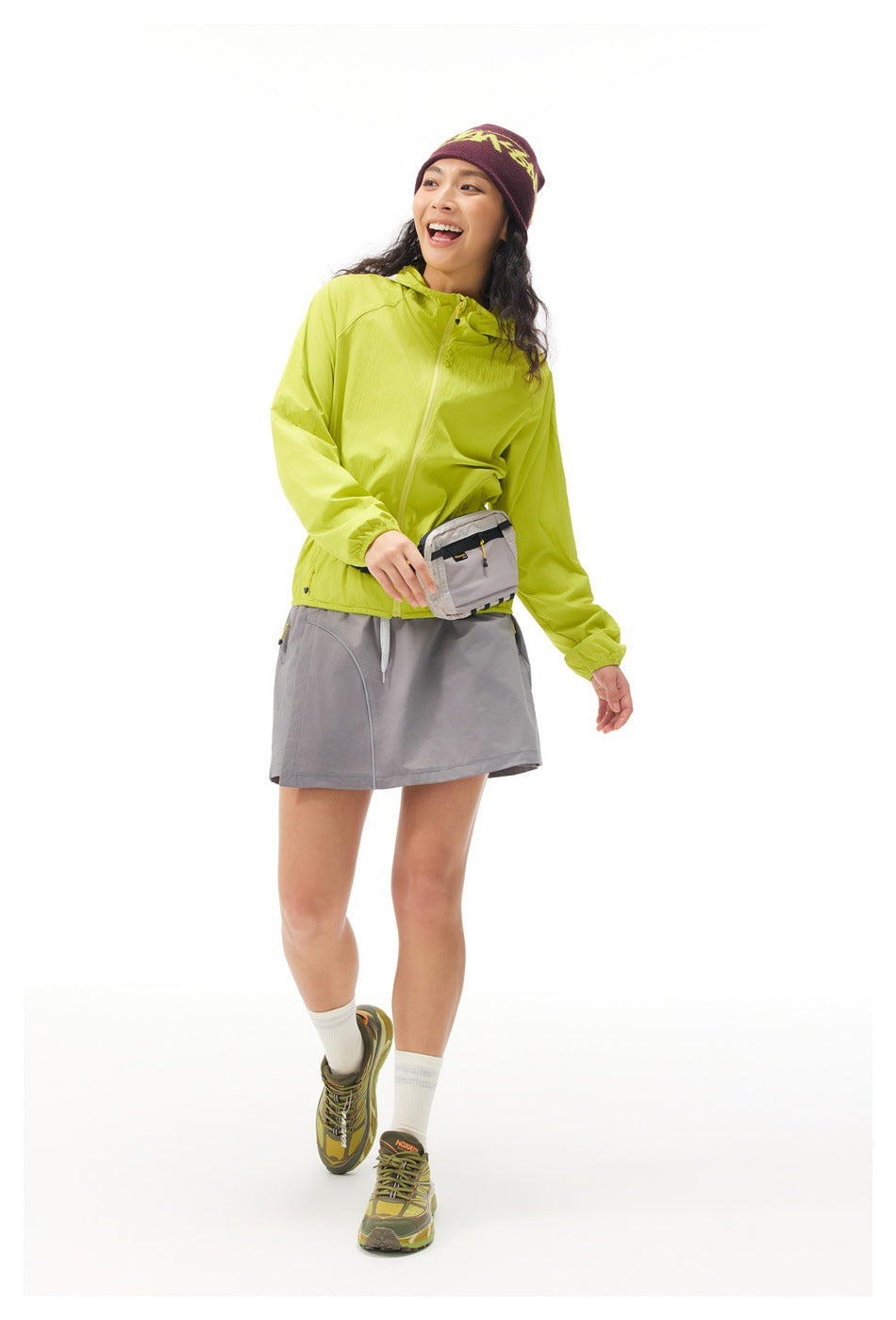 Full-length model wearing a lime hooded jacket, gray skirt, waist pack, and burgundy beanie with yellow lettering.