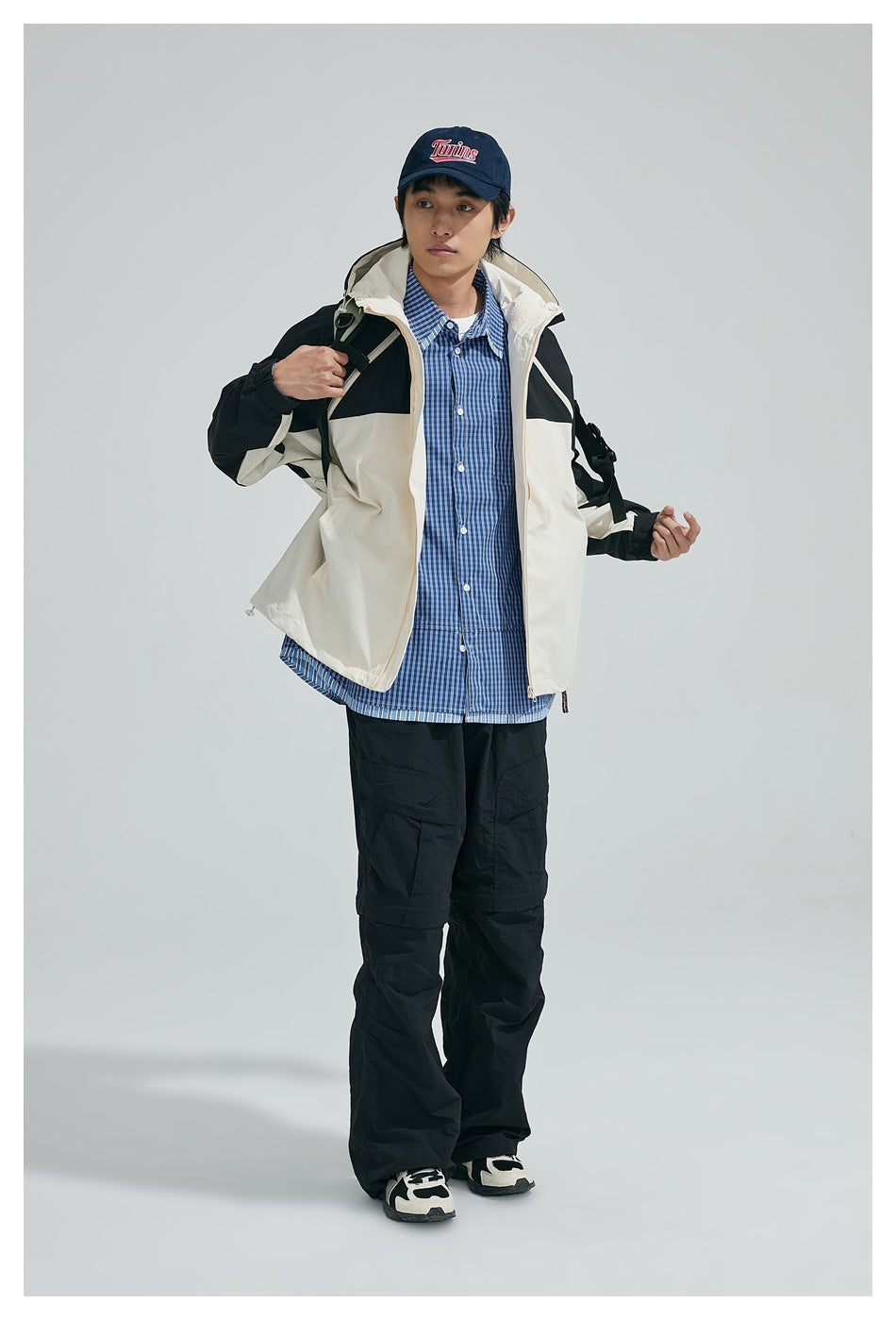 Model in black and beige hooded jacket with a backpack strap over a blue checked shirt, turned three-quarter pose