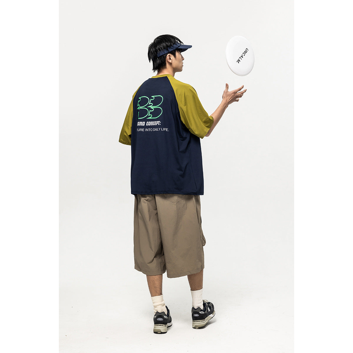 Model shown from behind wearing a navy and olive raglan T-shirt with a large back graphic, tossing a white disc.