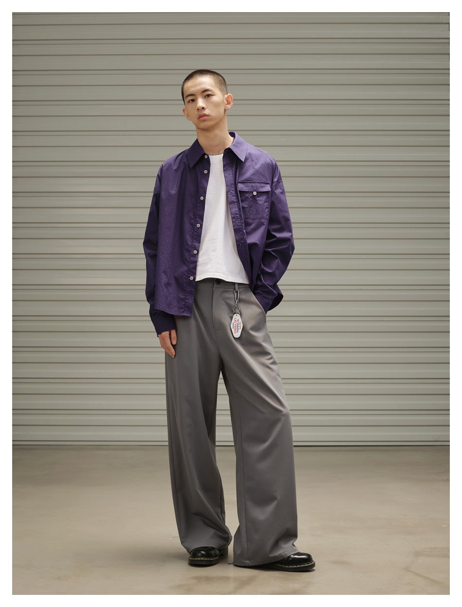 Model wearing a purple button-up overshirt, white T-shirt, and wide gray trousers, standing with one leg angled outward.