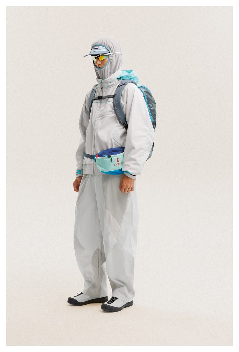 Model wearing a light gray hooded jacket and pants with a turquoise backpack and waist pouch, shown in profile.