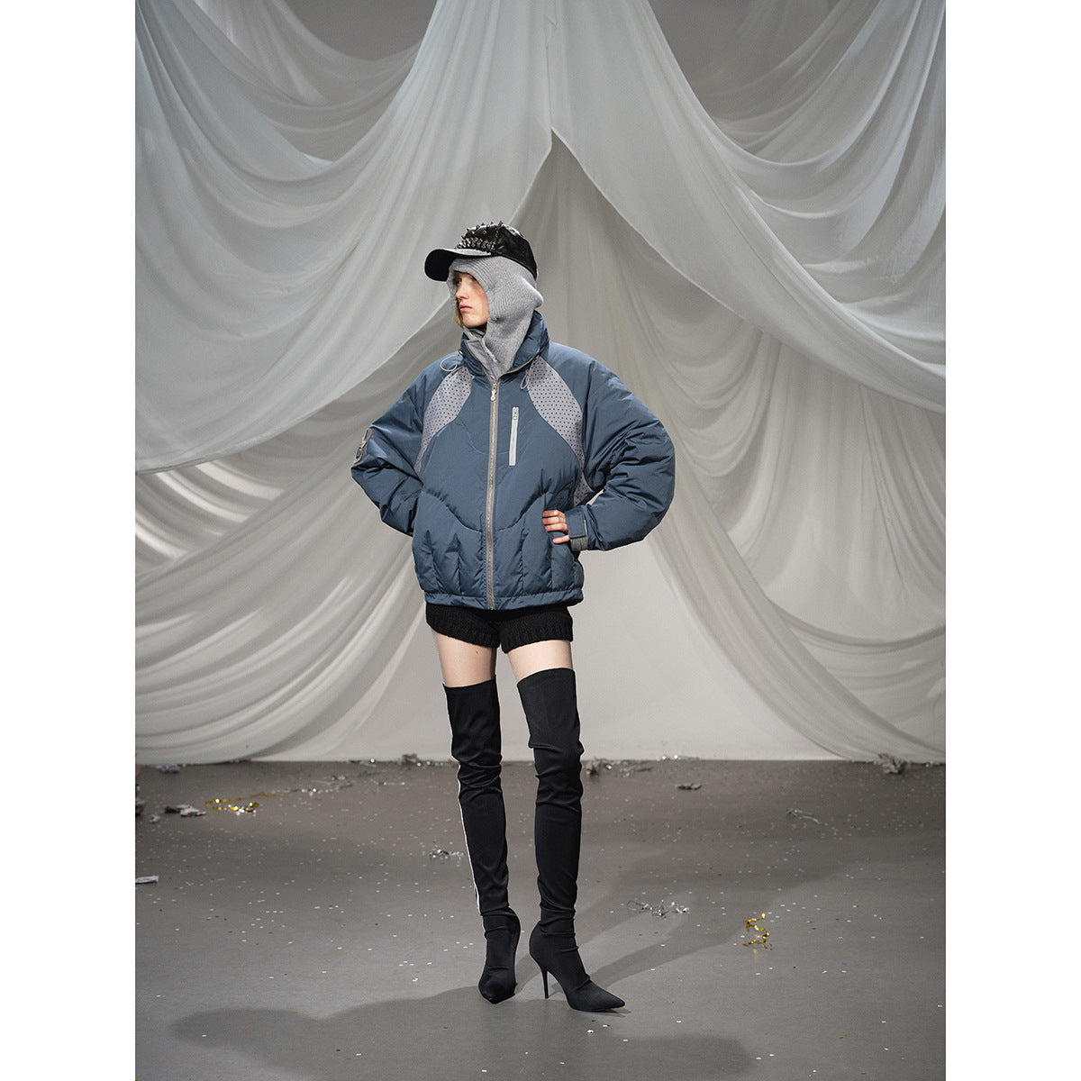 Full-length model wearing a blue-gray padded jacket with dotted shoulder panels, styled with black shorts and tall boots.