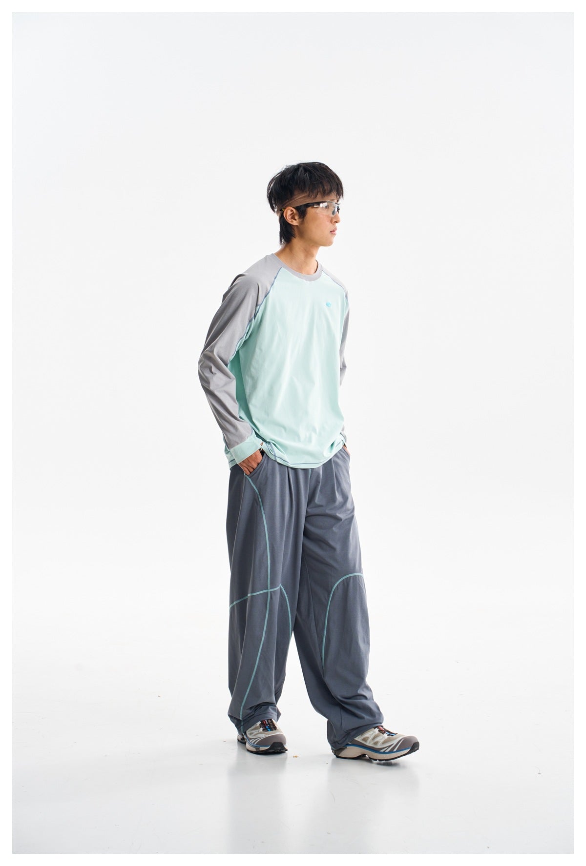 Full-length side view of a model wearing a mint-green and gray long-sleeve shirt, loose slate-blue trousers, and sneakers.