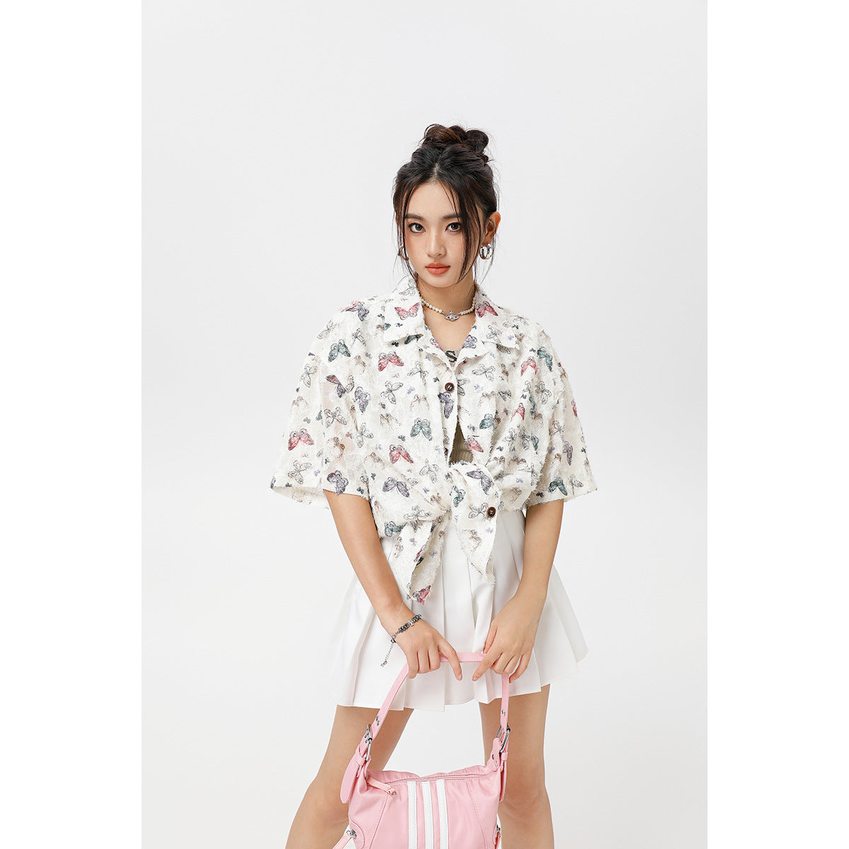 Model wearing a white short-sleeve button-up shirt with a multicolor butterfly print, tied at the waist over a pleated skirt.