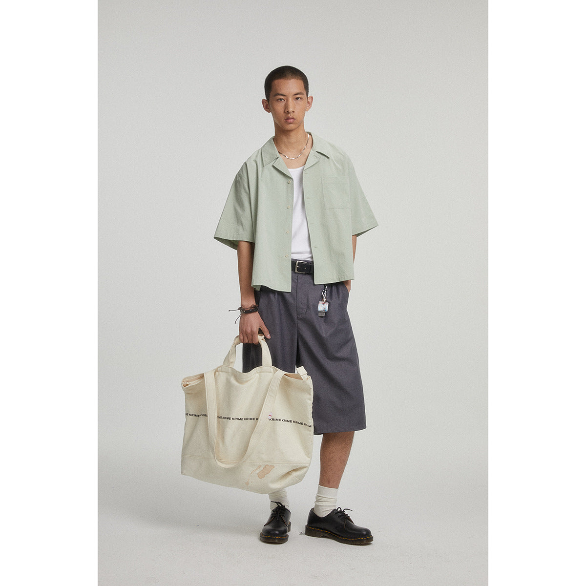 Model in a pale sage short-sleeve button-up over a white top with charcoal shorts, carrying a large cream tote.