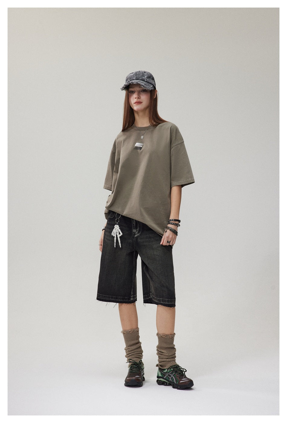 Full-length model wearing an oversized olive-gray T-shirt with a small chest graphic and black denim shorts.