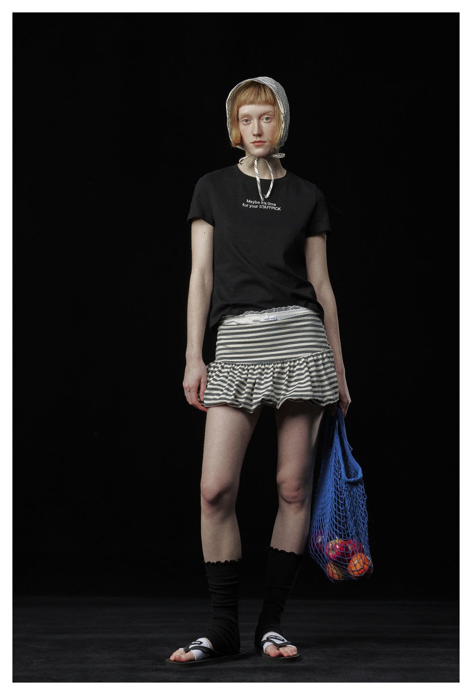 Model wearing a black short-sleeve slogan T-shirt with a striped ruffle mini skirt and bonnet, facing front
