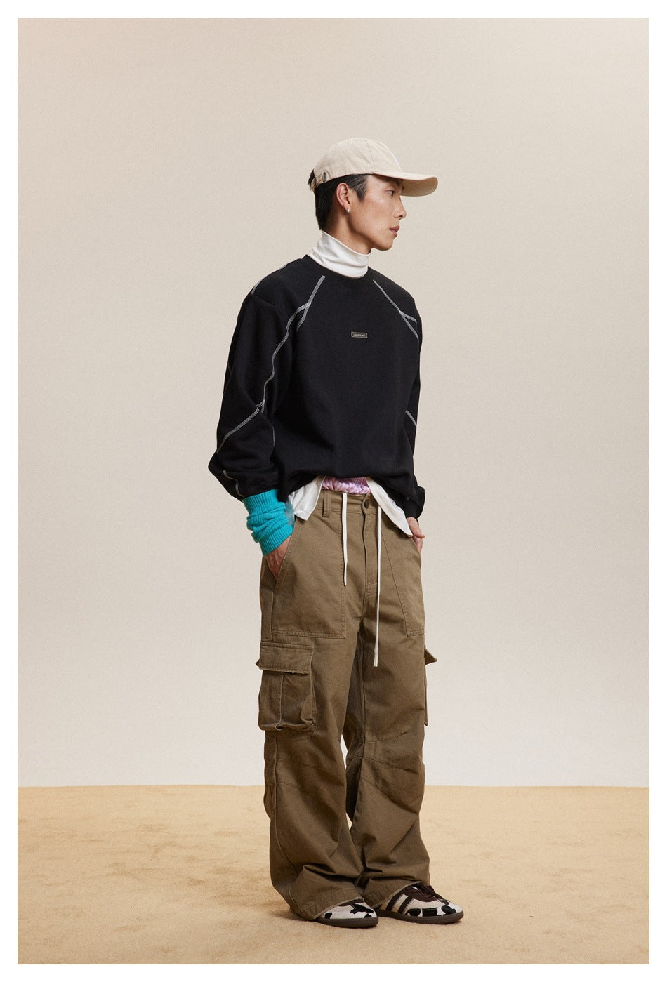 Side view of model in a black topstitched crew-neck sweatshirt with white turtleneck, khaki cargo pants and cap