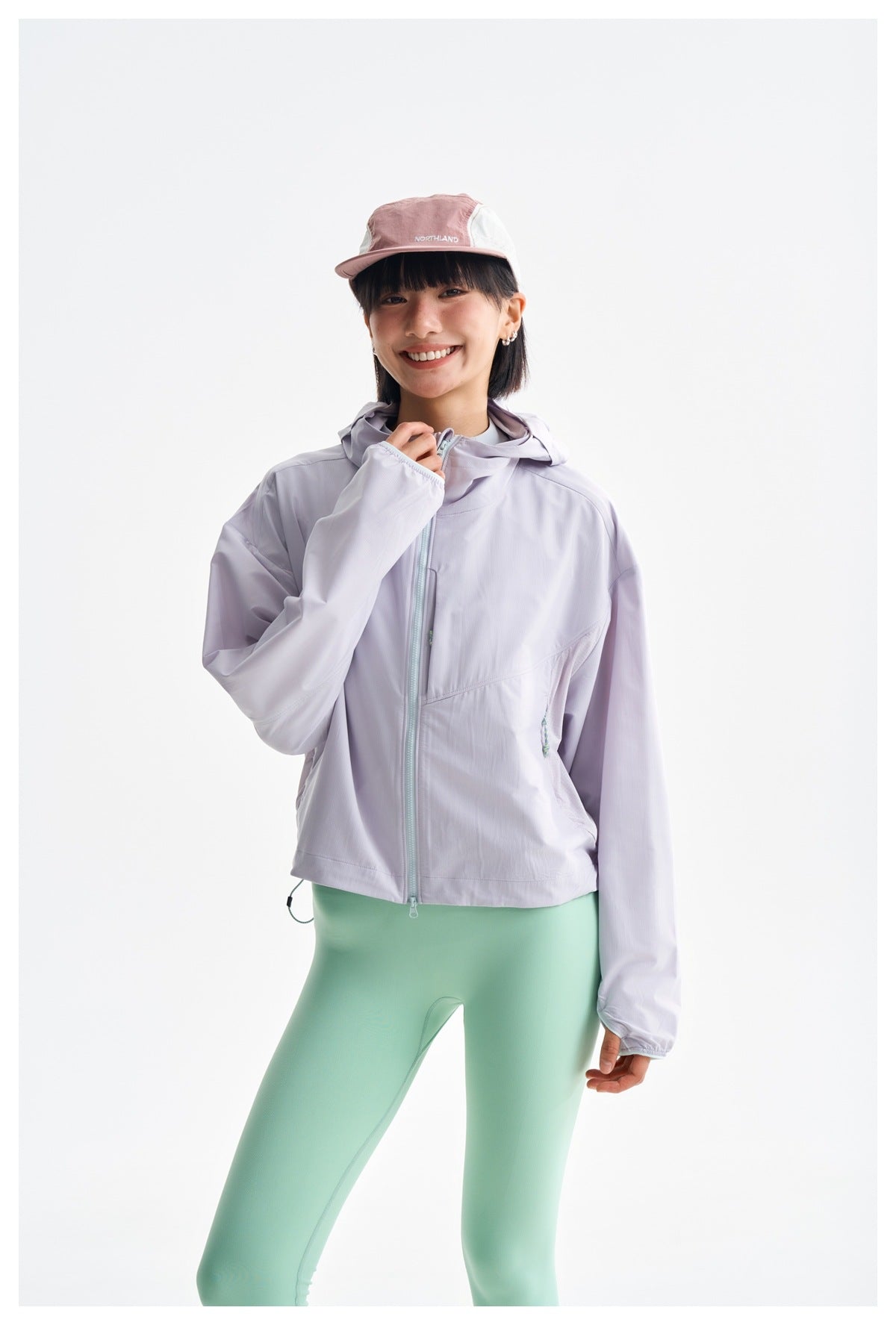 Model wearing a light lavender zip-front hooded jacket with mint leggings and a pink-and-white cap.