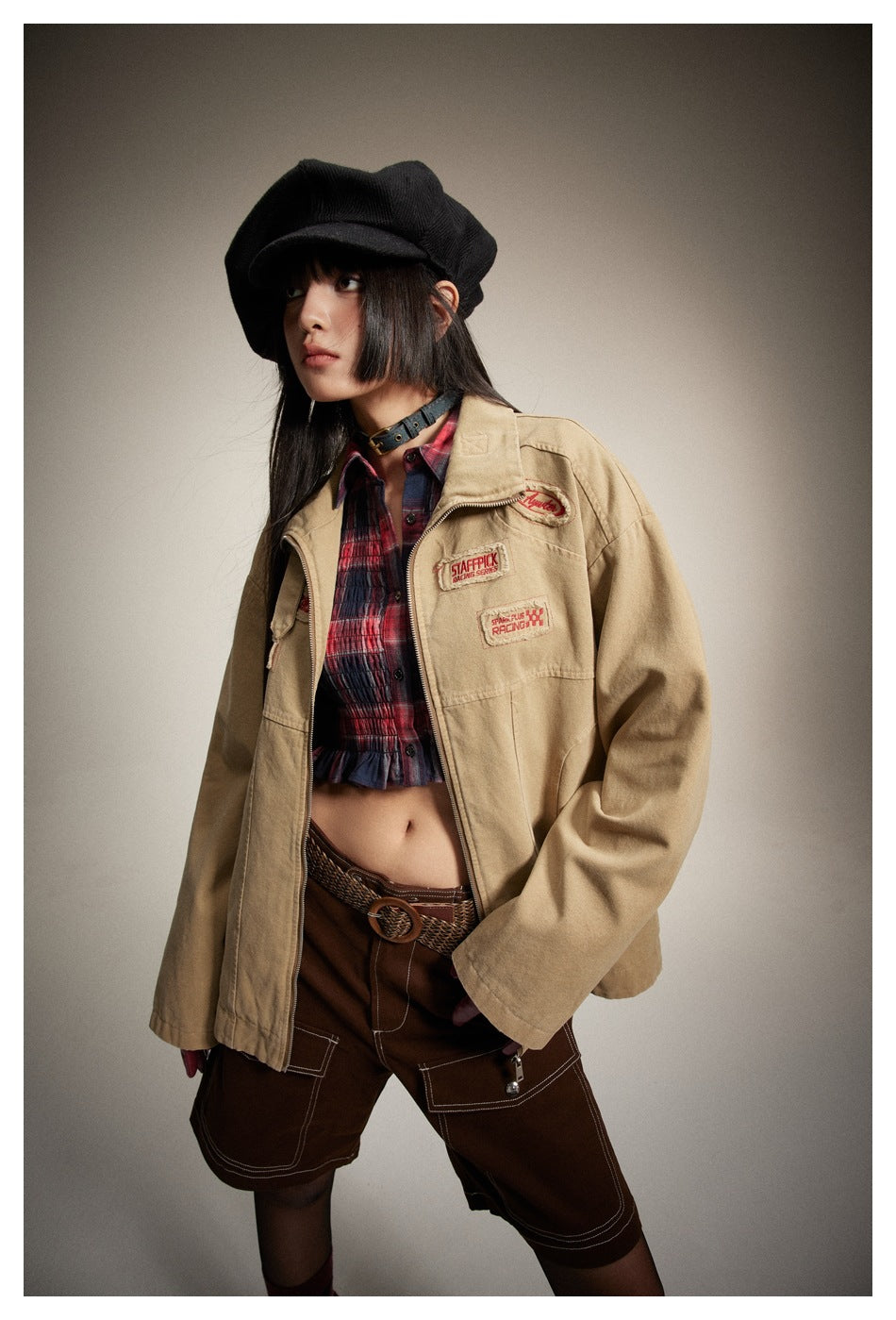 Model wearing a tan patch-decorated jacket open over a red plaid crop top and brown cargo shorts.