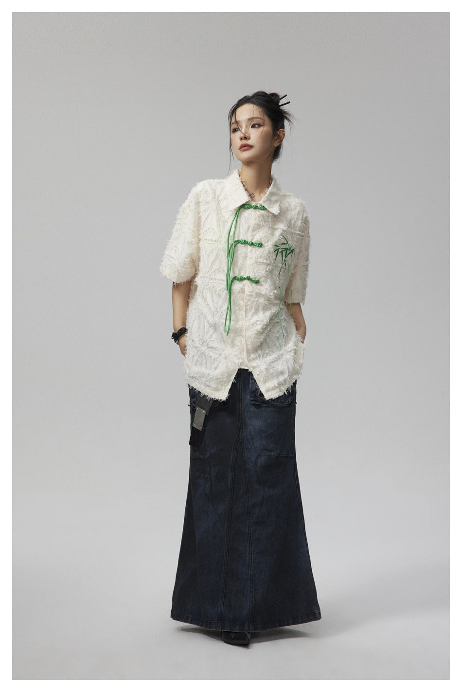 Model wearing an ivory textured short-sleeve jacket with green frog closures and a dark denim maxi skirt.