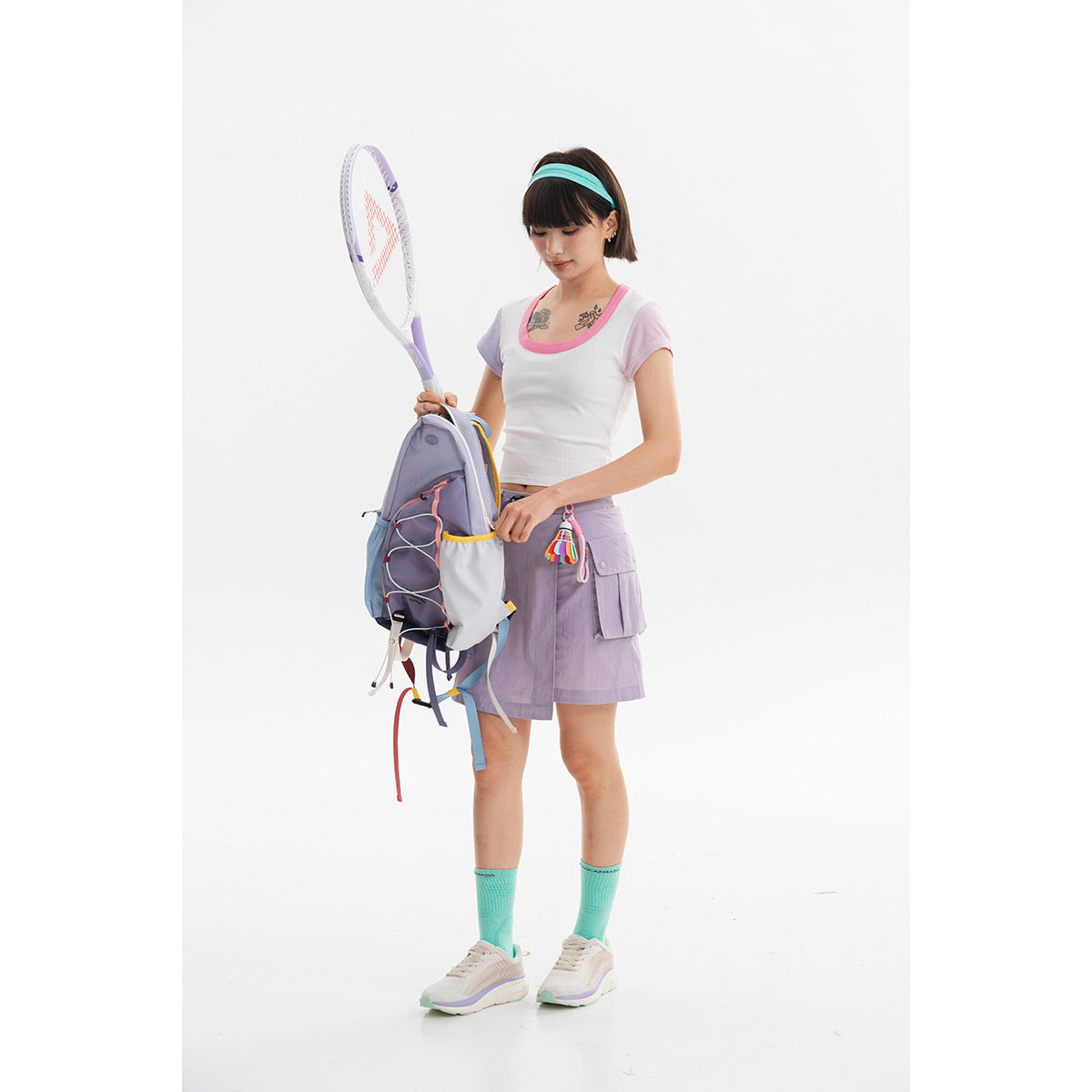 Model wearing a white cropped top with pink trim and lilac shorts, holding a tennis racket beside a backpack.