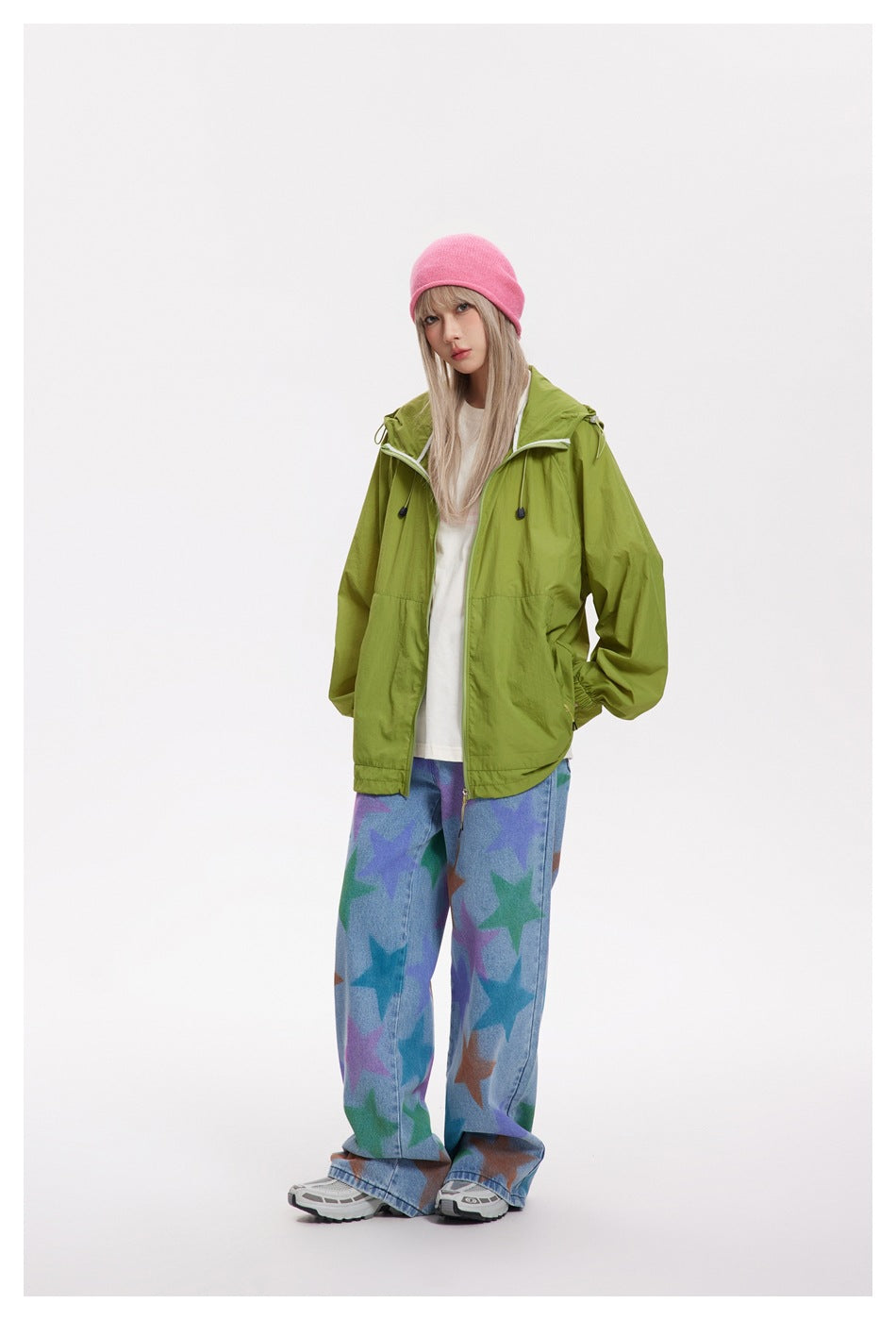 Model wearing green hooded zip jacket open over a white tee with star-print jeans and a pink beanie, front view
