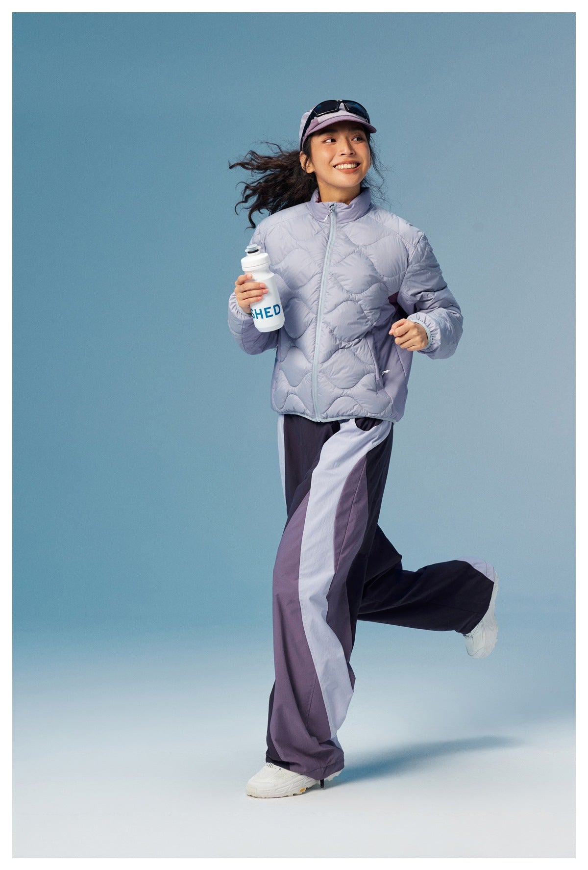 Woman in a pale lavender wave-quilted zip jacket and dark color-block wide-leg pants, posed mid-stride.