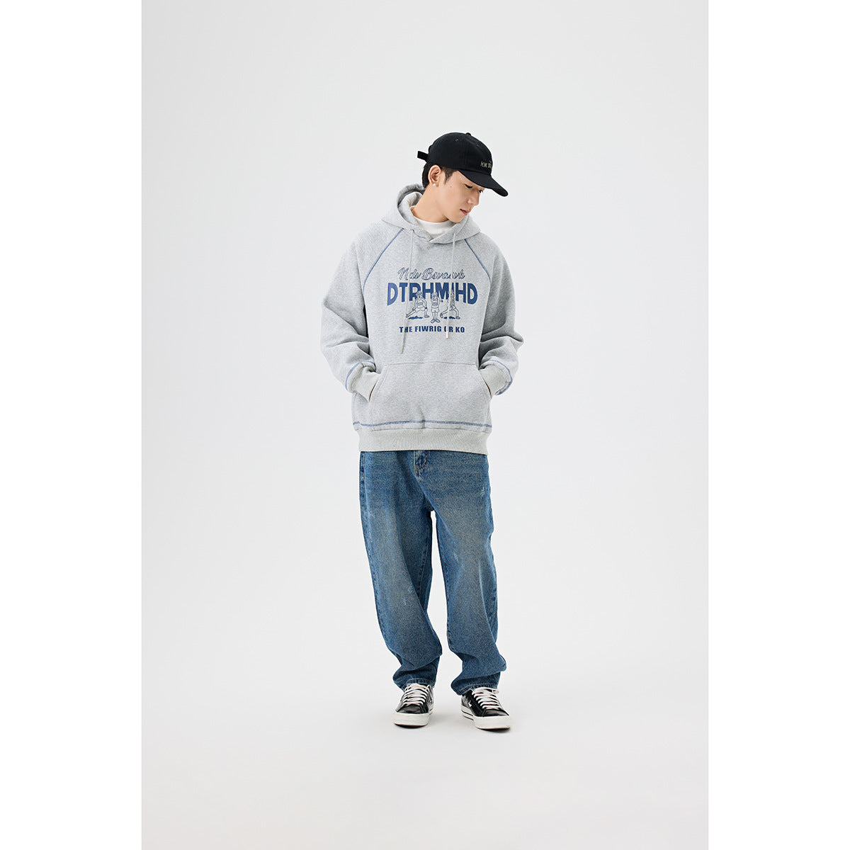 Model in a light gray graphic hoodie and loose blue jeans, shown standing with hands in the kangaroo pocket.