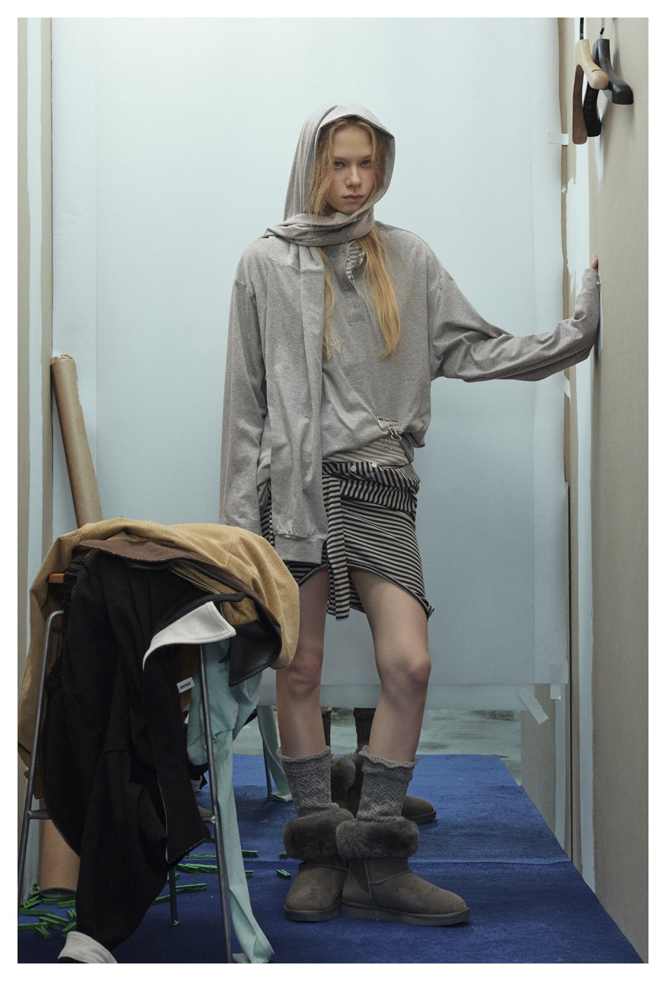 Model leaning on a wall in a gray long-sleeve henley top with a hooded shoulder layer, striped skirt layer and boots