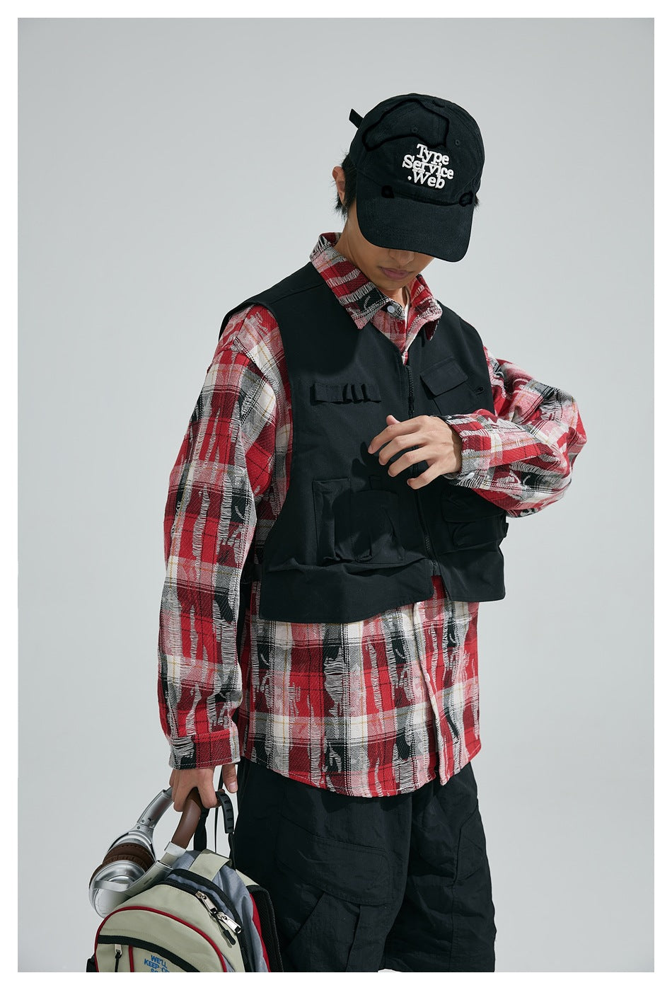 Model wearing a black utility vest over a red plaid shirt, black cap and trousers.