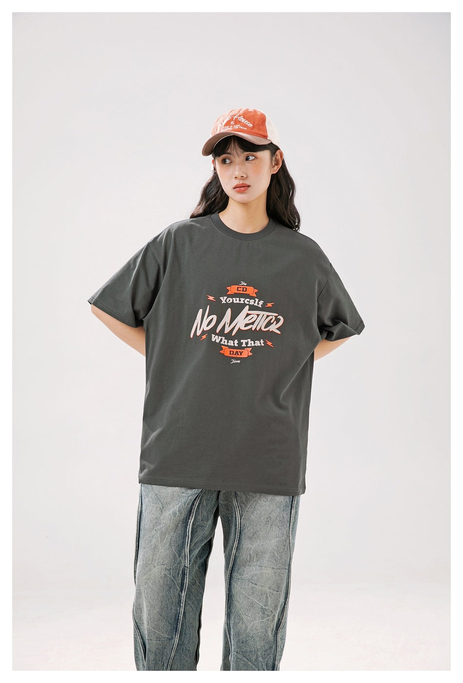 Person wearing a dark gray graphic T-shirt with orange script print, an orange cap and washed jeans, front view