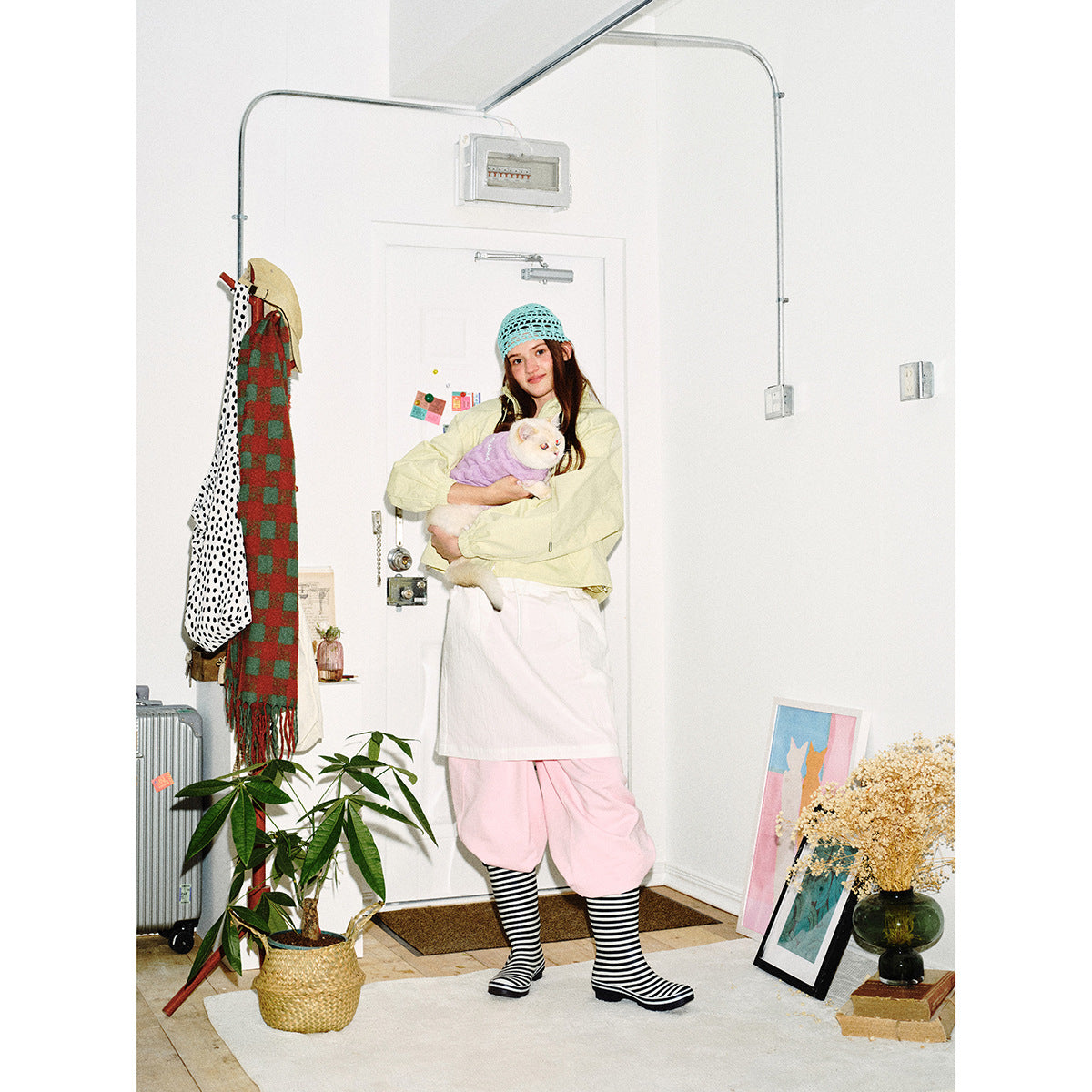 Full-length model wearing a pale yellow jacket and pink trousers, holding a cat in a purple sweater.