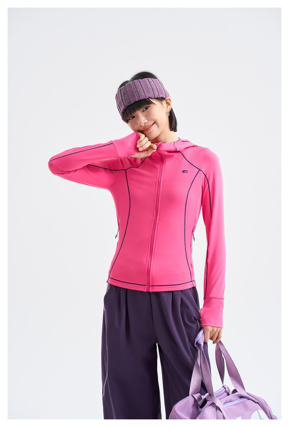Model in a bright pink hooded zip jacket and loose purple pants, posing with one sleeve raised.
