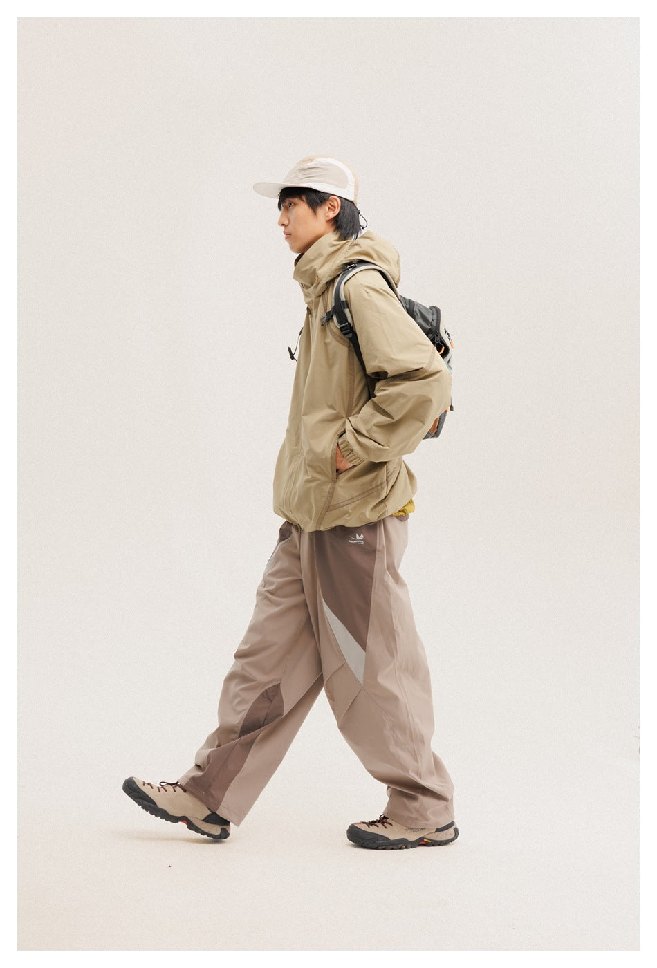 Model in a tan hooded jacket and loose taupe color-block pants, carrying a dark backpack.