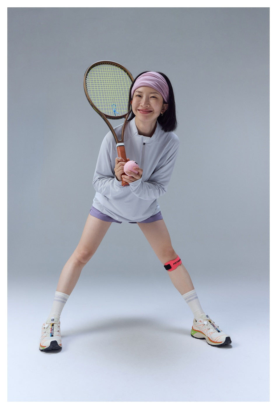 Model in a pale gray quarter-zip top and lavender shorts, crouching in a tennis stance with racket and ball.