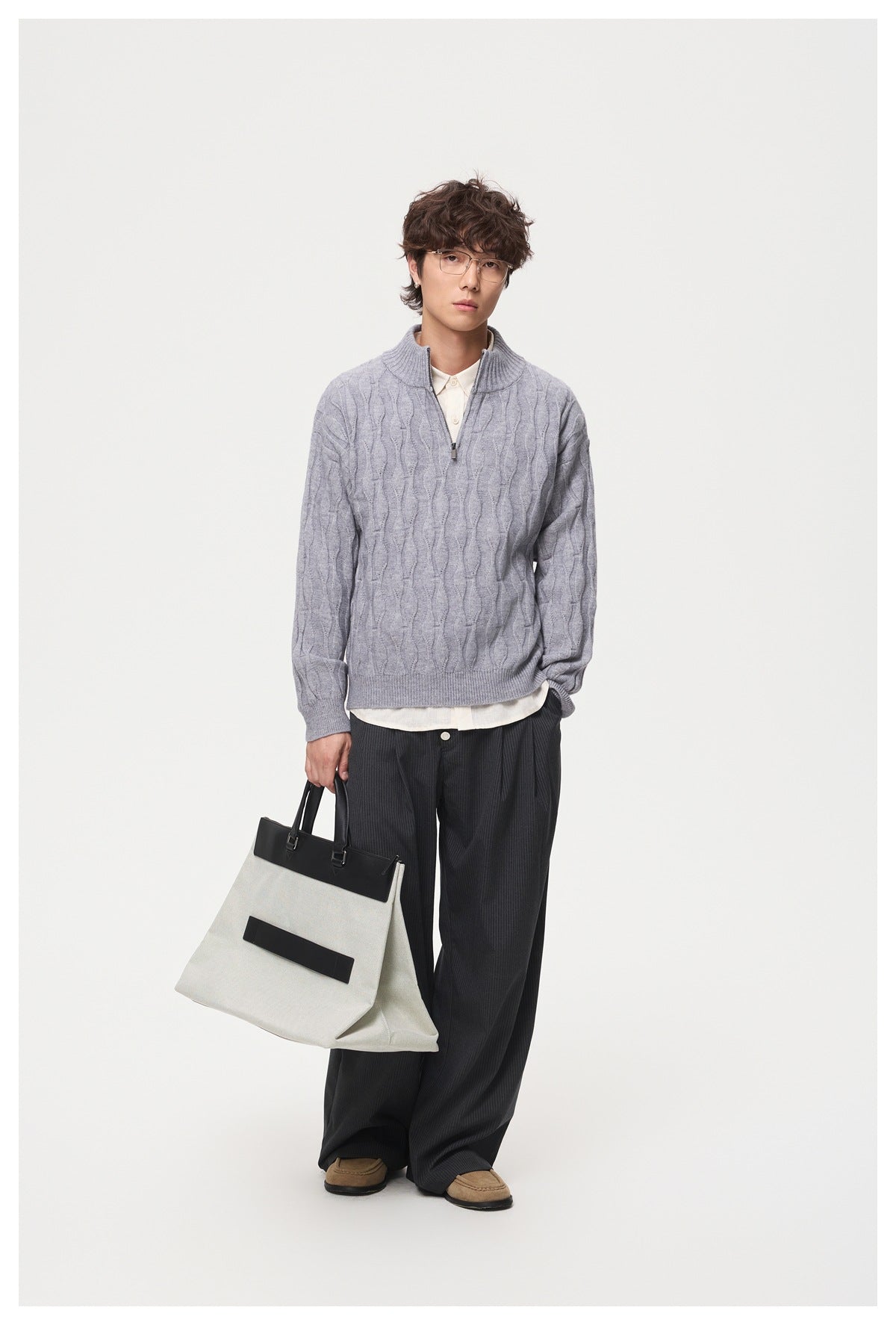Model wearing gray cable-knit half-zip sweater over a cream shirt with dark pinstripe trousers, holding a tote bag