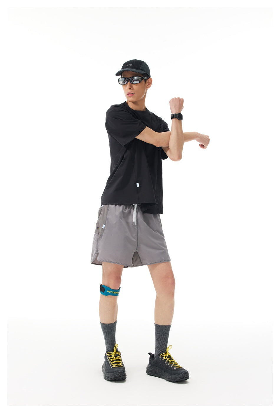 Model wearing a black short-sleeve shirt and gray athletic shorts, stretching one arm.