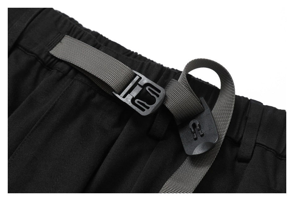 Close-up of black trousers showing a gray webbing belt and plastic buckle at the elastic waistband.