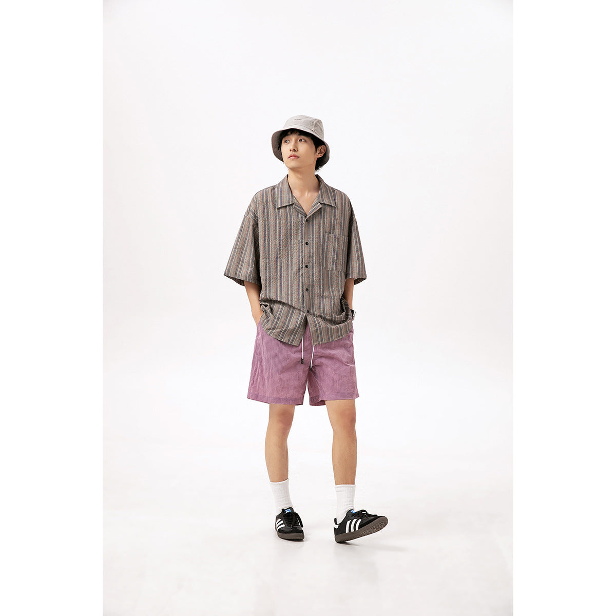 Model wearing a loose gray-brown striped short-sleeve button-up shirt, mauve shorts, and a light bucket hat.