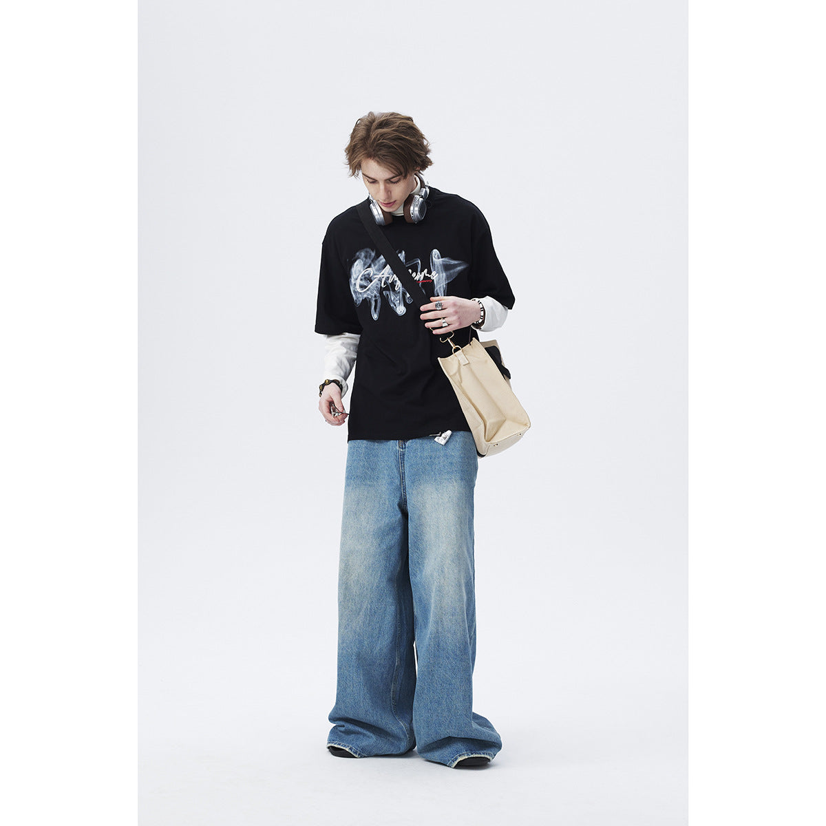 Model wearing a black graphic T-shirt over white sleeves with wide-leg blue jeans and a beige shoulder bag.