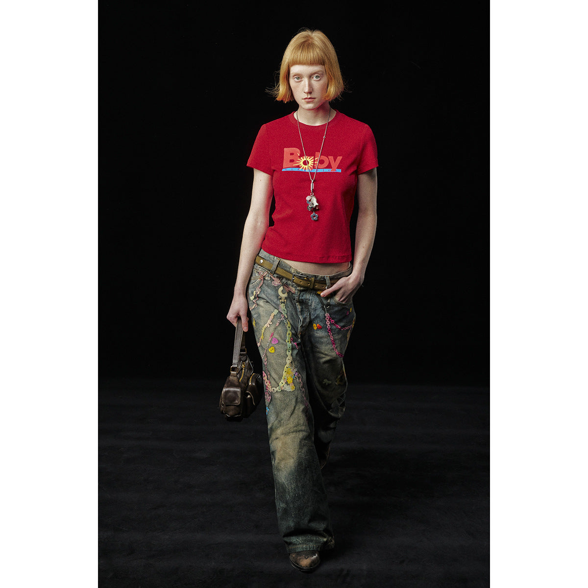 Full-length model wearing a red graphic T-shirt and decorated wide-leg jeans against a black background.