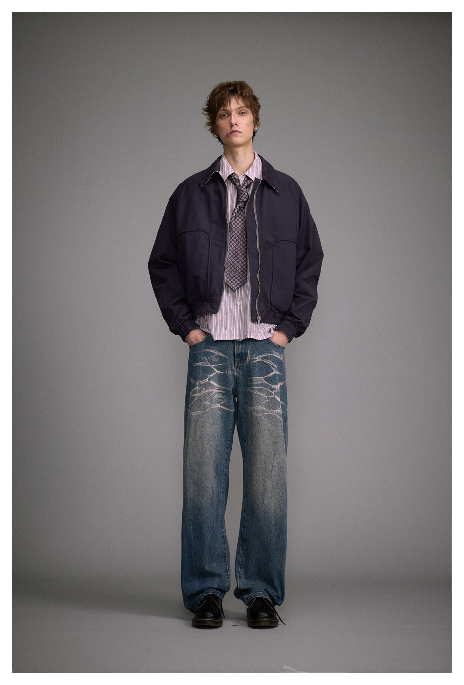 Model standing front view wearing wide-leg washed blue jeans with a dark jacket, striped shirt, tie and black boots