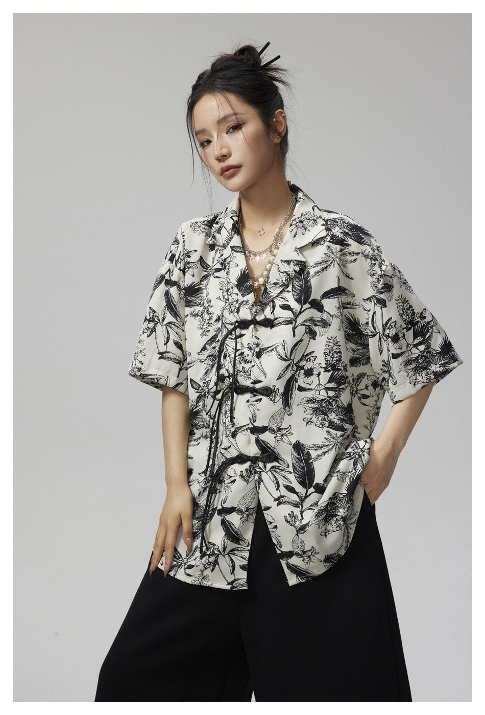 Model wearing an ivory botanical-print short-sleeve shirt with black knotted closures and wide black trousers, front view.