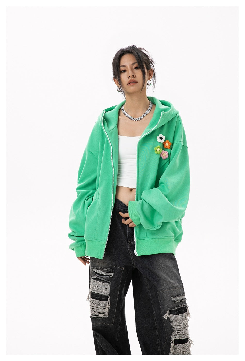 Model wearing an open green zip hoodie with flower appliques over a white crop top and ripped black jeans