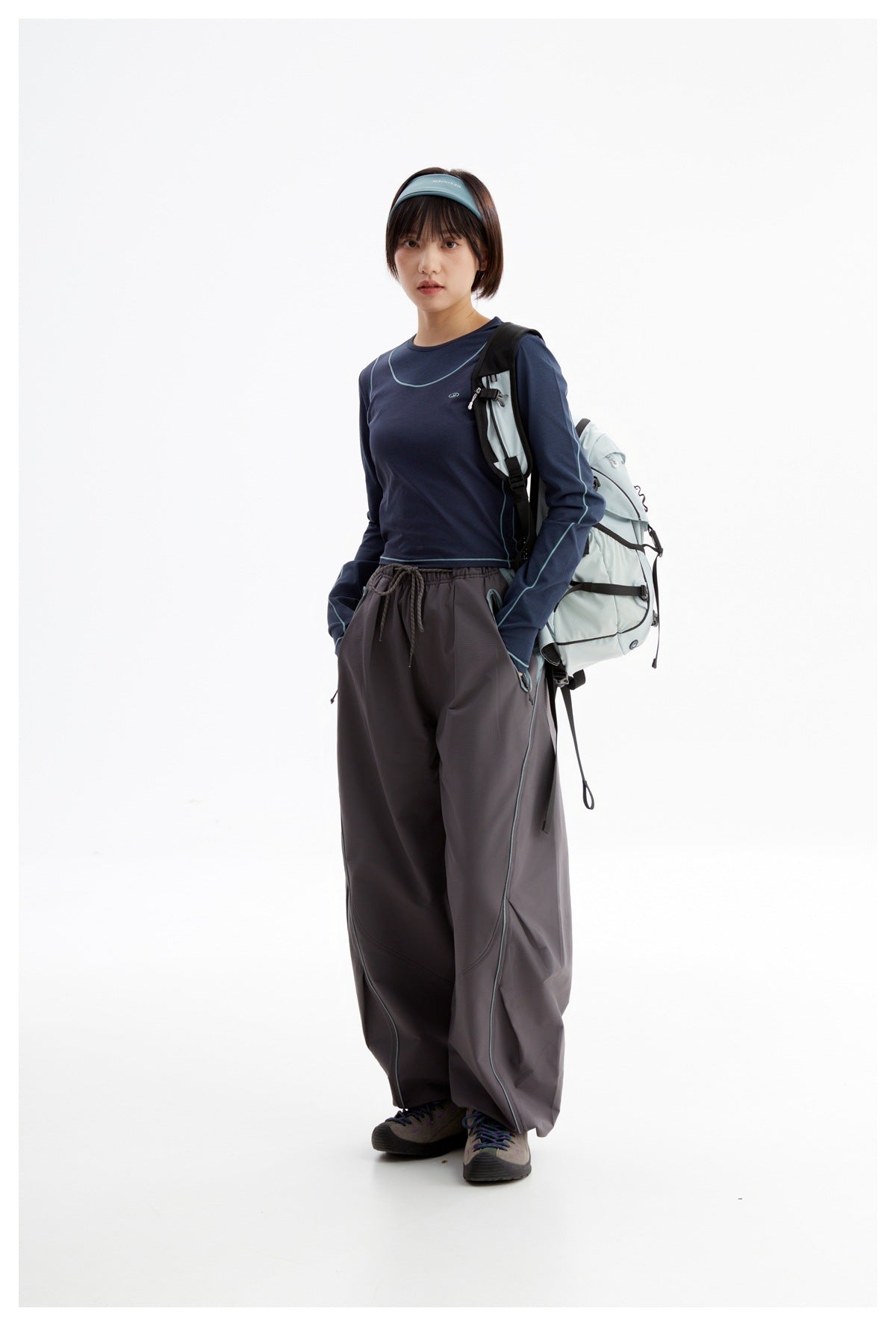 Model wearing a navy top with turquoise piping and charcoal wide-leg pants, carrying a pale backpack.