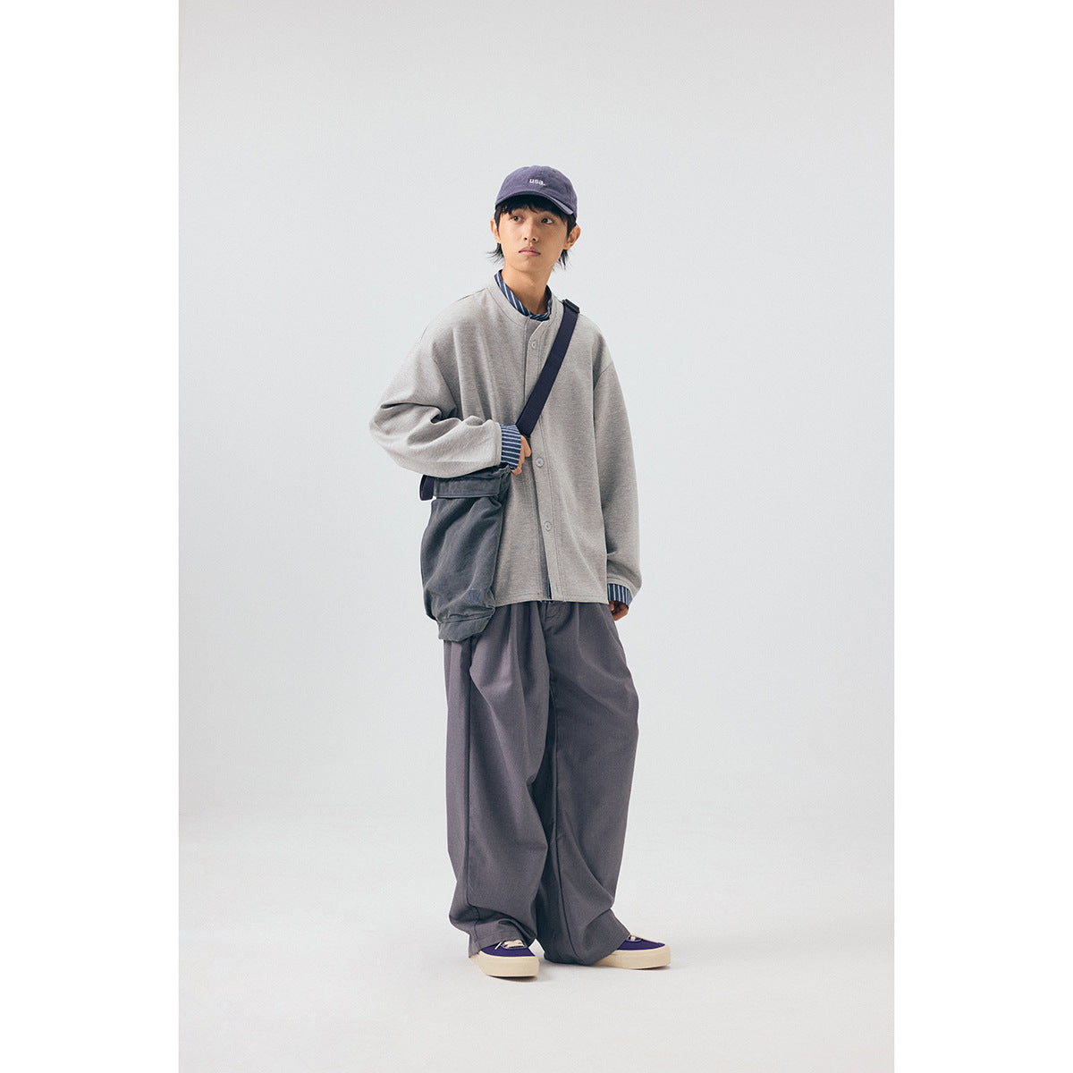 Model wearing a light gray button-front jacket with striped cuffs, loose charcoal trousers, and a crossbody bag.