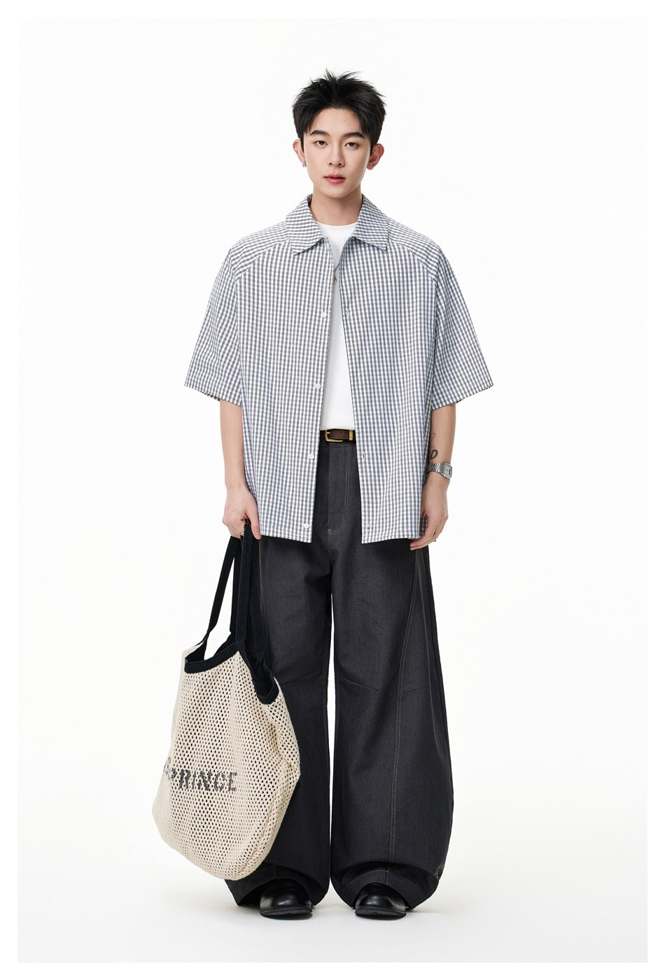 Model wearing an open check short-sleeve shirt over a white tee with wide dark pants and a mesh tote bag