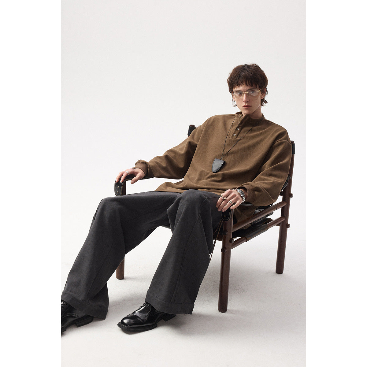 Model seated in a brown long-sleeve henley and wide charcoal trousers.